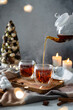Pouring hot tea into a glass cup in cozy winter composition. Two cups of black tea with steam and teapot in christmas composition with candles and christmas tree. Winter tea party, menu, poster