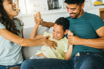 A happy family is spending their time together at home. The father is holding a boy and the mother is tickling him.