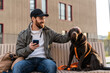 © NFstock - Overjoyed bearded man stroking his lovely dog and looking at him with toothy smile while using his smartphone at the bench during the walking