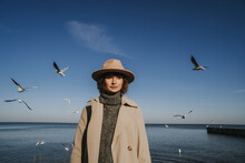 Woman In Overcoat And Hat Standing In Front Of Seagulls Flying Over Sea