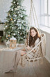 © Dina - Beautiful young woman on a boho style swing near christmas tree in the living room at home, decorated for winter holiday season.