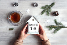 Calendar For January 16: Hands Hold A Decorative House With The Name Of The Month And The Number 16, A Cup With Hot Tea, Snowflakes, Fir Branches On A Gray Background, Top View