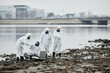 © Seventyfour - Wide angle view at group of people wearing hazmat suits collecting probes by water, toxic waste and pollution concept, copy space