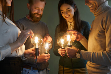 Team of people developing and trying out innovative ideas. Group of four clever men and women holding bright, shining, glowing Edison light bulbs to illuminate concept of creative idea and innovation