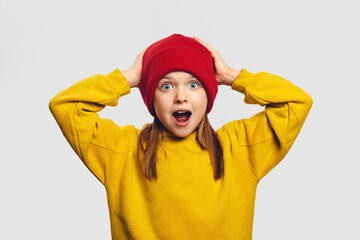 Portrait of shocked stupefied little kid in yellow hoodie looks with widely opened mouth, keeps hands on head, isolated over white background 