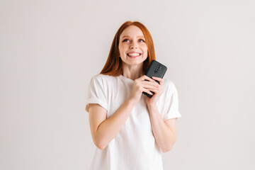 Studio portrait of happy dreaming young woman with wide smile reading good online message using mobile phone on white isolated background. Cheerful lady holding smartphone in hands and looking up.