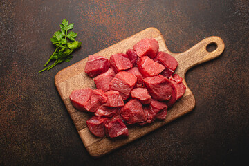 Wall Mural - Raw beef meat chopped in cubes with bunch of fresh parsley, garlic, salt and pepper on wooden cutting board for cooking stew or meat dish on brown dark stone concrete background top view flat lay