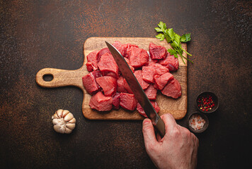 Wall Mural - Top view of a male hand chopping raw beef meat in cubes on wooden cutting board for cooking stew or other meat dish on brown dark stone concrete background overhead flat lay food concept 