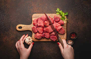 Wall Mural - Top view of a female hands chopping raw beef meat in cubes on wooden cutting board for cooking stew or other meat dish on brown dark stone concrete background overhead flat lay food concept