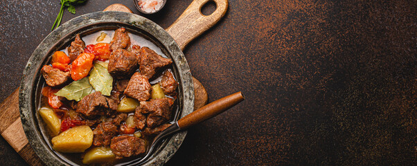 Delicious beef meat stew dish with potatoes, carrot and gravy in rustic metal bowl with spoon, bunch of fresh parsley, garlic cloves, spices on brown concrete background top view space for text