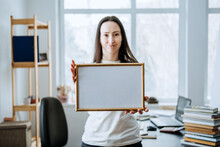 Frame Picture, Poster, Diploma, Certificate Mockup In Female Hands. Young Brunette Woman Holding Empty Blank Wooden Photo Frame On Table With Laptop And Books Home Office Background