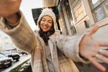 Happy Young Caucasian Lady Smiles With All Her Might And Reaches Out To Hug Camera Against Background Of Street. Beauty With Dark Hair Wearing Hat, Sweater And Coat. Winter Fashion, Holidays Concept.