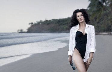 Wall Mural - A stylish sexy curly haired lady dressed in a bathing suit and a white shirt walks along the beach