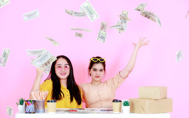 Portrait studio shot of two Asian happy rich wealthy female startup small business entrepreneur businesswoman partner sitting together throwing hundred dollar banknotes raining on pink background