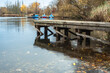 In the foreground a jetty protrudes into the lake, in which autumnal leaves swim. Boats are attached to the jetty on the other bank.