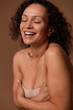 Delighted dark-haired curly woman with closed eyes and skin flaws and imperfections in beige underwear smiles toothy smile posing against beige background with copy space for ads