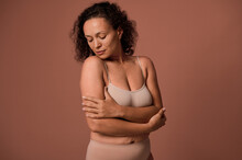 Confident Serene Beautiful Curly Woman With Imperfect Body, Cellulite And Stretch Marks After Childbirth, In Beige Underwear, Looking Down, Hugging Herself On A Colored Background With Copy Ad Space