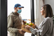 © Syda Productions - food shipping, pandemic and people concept - delivery man in mask giving order in wooden box to female customer at home