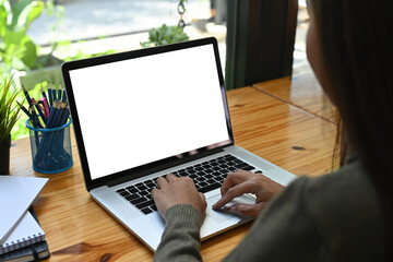 behind view of a young woman using a white blank screen computer laptop at the wooden working desk.