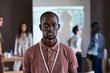 © AnnaStills - Portrait of African young businessman looking at camera while standing in the boardroom with other people in the background