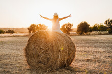 Carefree Woman With Arms Outstretched Sitting On Straw Bale In Field