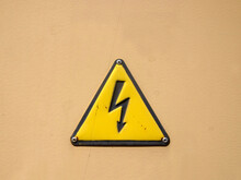 High Voltage Dangerous Electric Sign. View Of A Triangular Yellow Warning Sign On A Beige Wall.