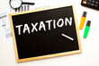 © Ihar - Text TAXATION written in chalk on a slate board. Office desk with office supplies.