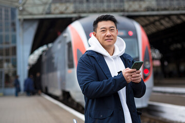 Wall Mural - Male Asian businessman, at the train station, buys train tickets online, uses a mobile phone and an application, the tourist returns home from a business trip