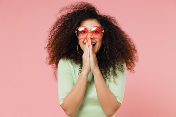 Poster - Overjoyed excited shocked surprised stupefied young curly latin woman 20s years old wears mint t-shirt sunglasses cover mouth with hand isolated on plain pastel light pink background studio portrait.