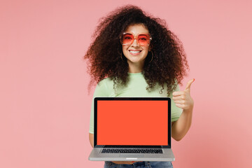 Wall Mural - Young curly latin woman 20s wear mint t-shirt sunglasses hold laptop pc computer with blank screen mock up showing thumb up like gesture isolated on plain pastel light pink background studio portrait