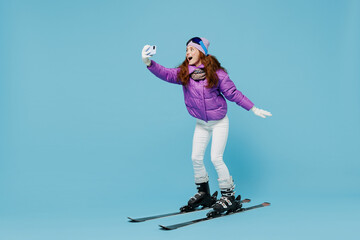 Wall Mural - Full body skier amazed fun woman in warm purple padded jacket ski goggles mask spend extreme weekend in mountains do selfie shot on mobile cell phone gesture isolated on plain blue background studio