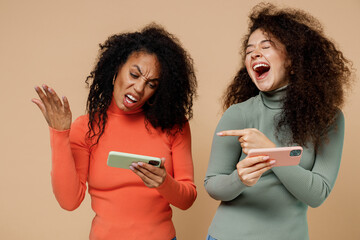 Poster - Two young curly black women friends 20s wear casual clothes play racing app on mobile cell phone one lose another point finger laughing isolated on plain pastel beige background studio portrait