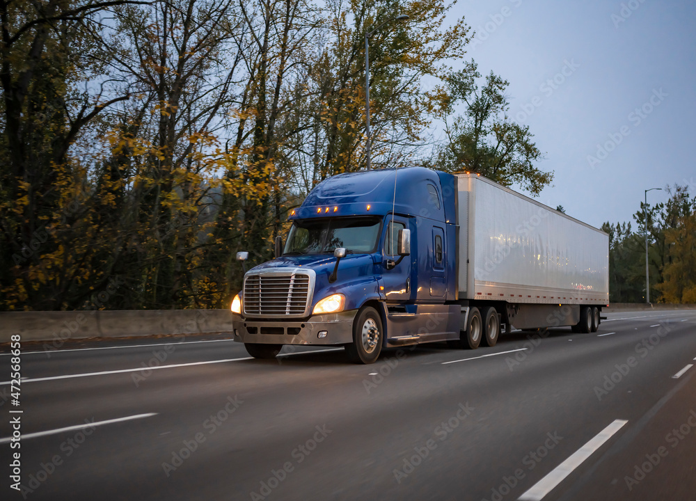 Dark blue big rig semi truck with turned on headlights transporting ...