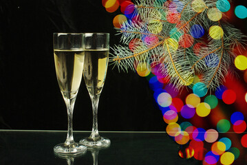 Canvas Print - Christmas evening. Two glasses of champagne, a branch of a fir tree and multi-colored glare on a dark background.