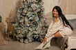 © Тамара Киреева - a beautiful dark-haired girl in a knitted sweater and knee socks is sitting on the bed by the Christmas tree