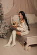 © Тамара Киреева - a beautiful dark-haired girl in a knitted sweater and knee socks is sitting on the bed by the Christmas tree with a gift box in her hands