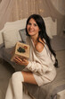 © Тамара Киреева - a beautiful dark-haired girl in a knitted sweater and knee socks is sitting on the bed by the Christmas tree with a gift box in her hands