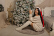 © Тамара Киреева - a beautiful dark-haired girl in a knitted sweater and knee socks is sitting by the bed at the Christmas tree in a Santa Claus hat in the hands of a mug
