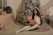 © Тамара Киреева - a beautiful dark-haired girl in a knitted sweater and knee socks is sitting by the bed at the Christmas tree in a Santa Claus hat in a gift box