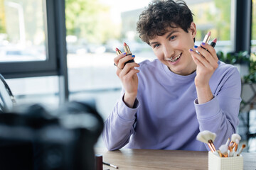 Positive transgender blogger holding lipsticks near blurred digital camera.