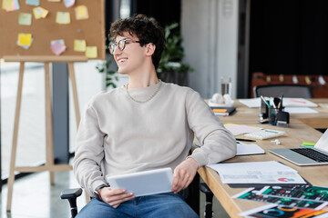 Wall Mural - Smiling transgender person holding digital tablet near papers with charts on table in office.