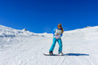 A girl snowboarder in ski goggles with loose hair stands alone on a ski slope. In the background snowy mountains and clear blue sky. Active winter holidays in the mountains.