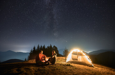 Wall Mural - Woman with guitar in her hands sitting opposite man by bonfire near their illuminated tent. Romantic evening outdoors under starry sky in the mountains. Concept of night camping and relationships.