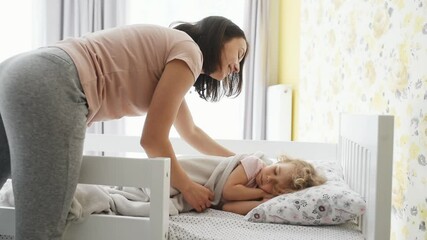 young mother kisses her little daughter that lying on the bed and going to sleep indoors at home