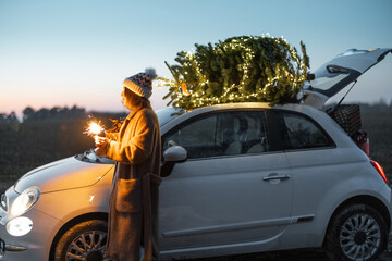 Wall Mural - Happy caucasian woman firing sparklers near car with Christmas tree on nature at dusk. Concept of celebrating New Year holidays. Idea of Christmas mood and fun. Side view with sunset sky on background