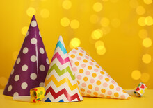 Beautiful Party Hats And Blowers On Yellow Table Against Blurred Festive Lights. Space For Text