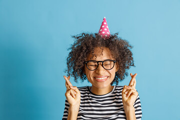 Wall Mural - Cheerful multiethnic lady in birthday cone hat keeping fingers crossed and smiling while making wish. Isolated on blue background