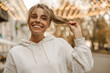 © Look! - Close-up of young caucasian blonde with gathered hair holding ends of her hair on blurred background. Smiling beauty in white sweatshirt smiles with teeth. Concept enjoying moments