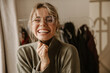 Close-up young caucasian woman laughing hard while screwing up eyes while being indoors. Girl with tousled blonde hair in sweater and glasses smiles broadly. Emotions people, lifestyle concept.