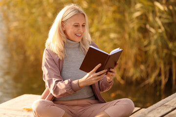 Sticker - Mature woman reading book near river on autumn day